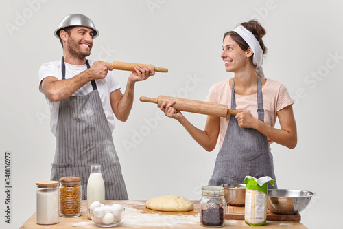 Horizontal shot of funny woman and man cook have culinary battle, pose at kitchen, shoot in each other with wooden rolling pins, have fun, make dough, prepare for Easter holiday, bake at home photo