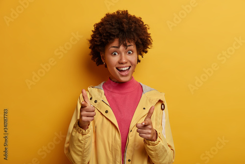 Hey you, attention. Clients choice concept. Positive African American girl points at camera with happy expression, chooses something, picks someone, wears casual clothes, isolated on yellow background