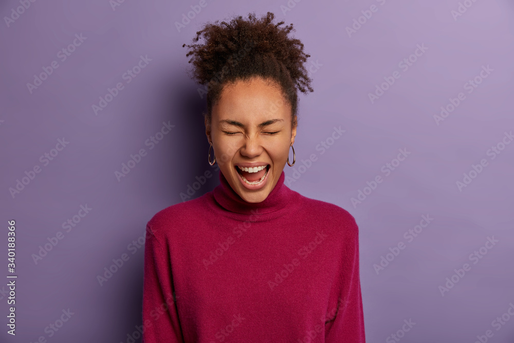 Happy dark skinned girl cannot hold laugh, squints face, giggles over funny joke, watches something adorable or funny, wears burgundy turtleneck, isolated on purple background, has positive day Happy dark skinned girl cannot hold laugh, squints face, giggles over funny joke, watches something adorable or funny, wears burgundy turtleneck, isolated on purple background, has positive day