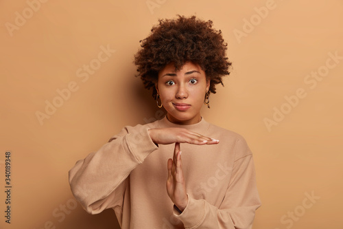 Photo of puzzled dark skinned woma makes time out gesture, has serious frustrated expression, curly hairstyle, dressed in casual sweatshirt, poses against beige studio background, needs to stop © wayhome.studio 