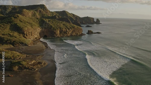 Aerial flying over calm West Coast ocean with waves crashing onto black sand rocky beach during West Coast sunset. Te Henga, Bethells Beach, Auckland, New Zealand. photo