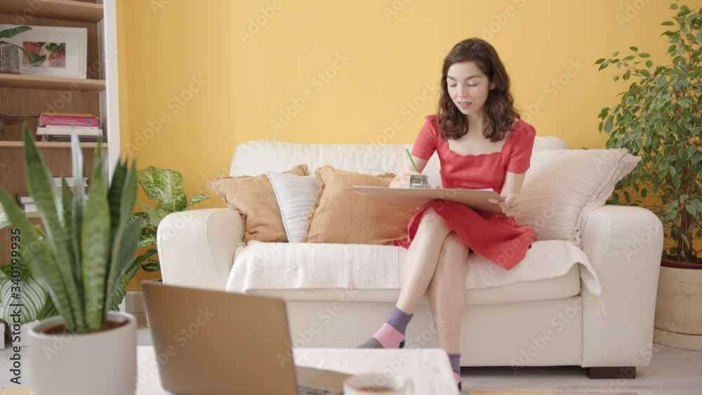 Portrait of a Young Beautiful Artist Girl with Braces. Attractive Woman Sitting on a Coach and Draws a Picture with Colored Pencils. Cozy Home interior, Yellow Wall, House Plants. Portrait of a Young Beautiful Artist Girl with Braces. Attractive Woman Sitting on a Coach and Draws a Picture with Colored Pencils. Cozy Home interior, Yellow Wall, House Plants.
