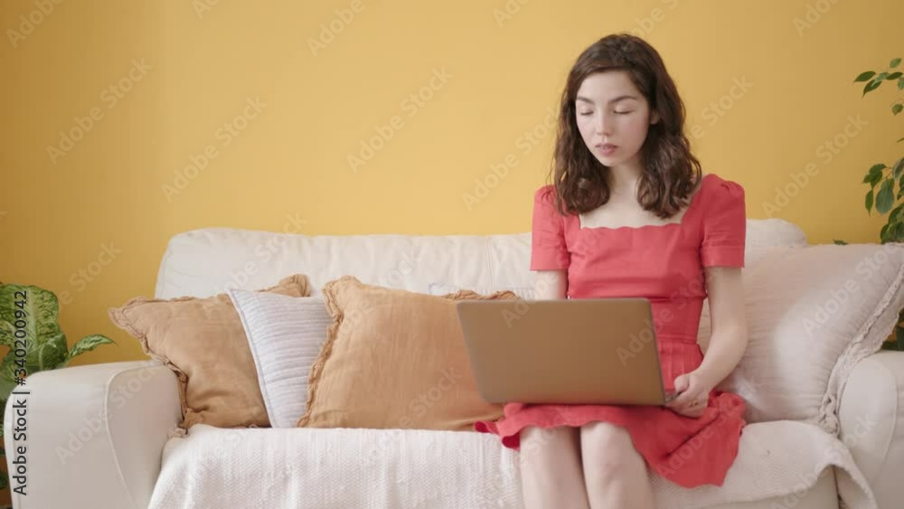 Young Attractive Woman in Red Dress Remotely Working with Laptop from Living Room. Cozy White Interior with Couch and House Plants. Concept of Home Office and Distant Work. Young Attractive Woman in Red Dress Remotely Working with Laptop from Living Room. Cozy White Interior with Couch and House Plants. Concept of Home Office and Distant Work.
