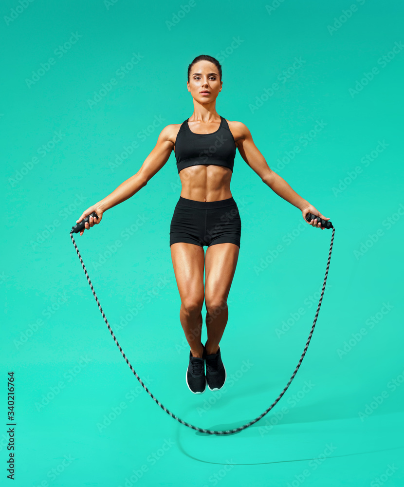 Sporty girl exercising with skipping rope. Photo of girl in black sportswear on turquoise background. Dynamic movement. Full length. Sports and healthy lifestyle - obrazy, fototapety, plakaty 