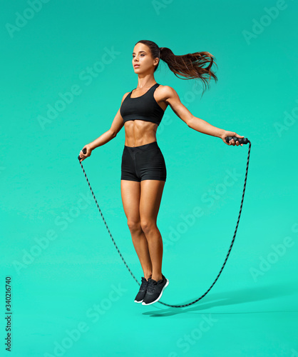 Sporty girl exercising with skipping rope. Photo of girl in black sportswear on turquoise background. Dynamic movement. Full length. Sports and healthy lifestyle