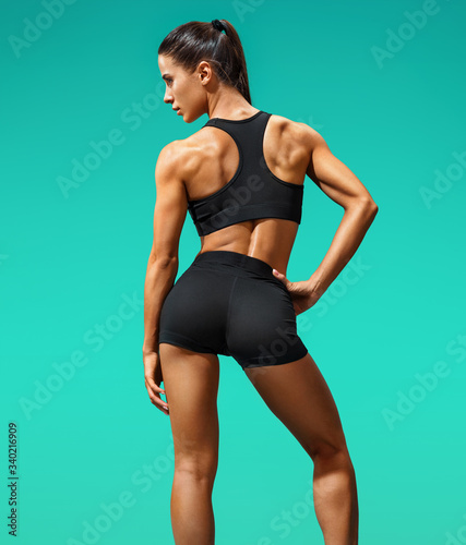 Sporty woman with slim tanned body in black sportswear posing on turquoise background. Rear view