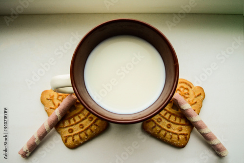 Closeup milk in a white and brown coffee mug  two gray-pink wafer rolls  two sugar cookies with patterns on the windowsill. on a white background. sugar products. sweet snack. inside. sweet pastries. 