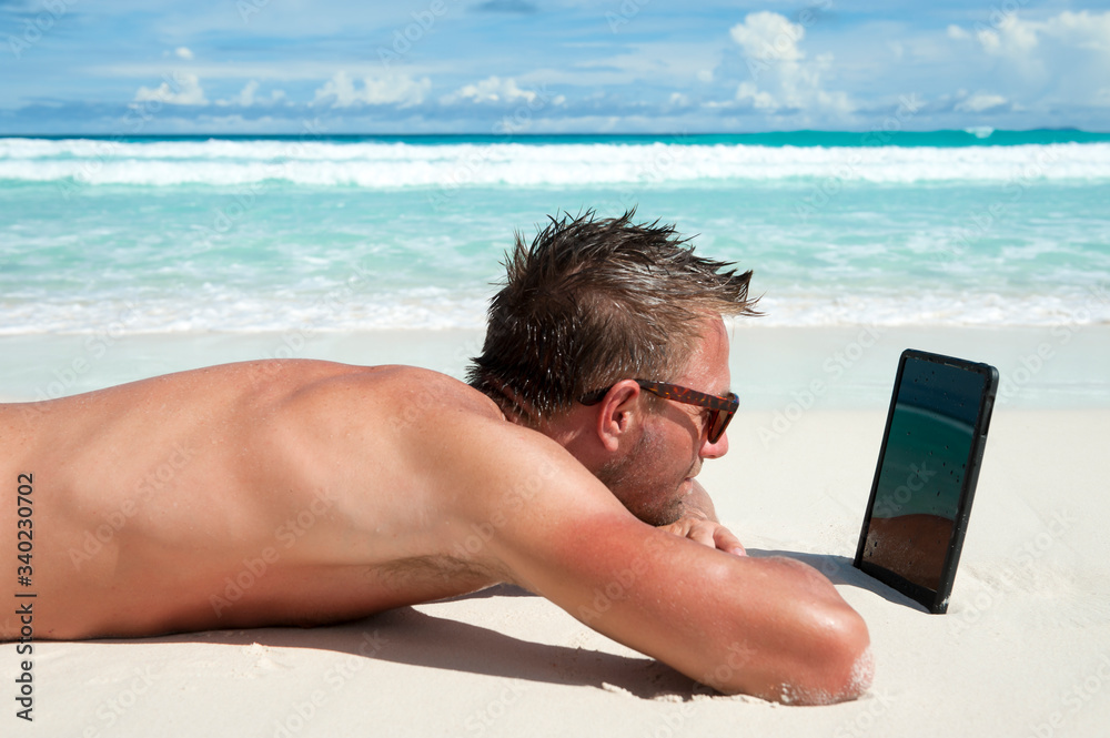 Man relaxing on tropical beach using digital tablet computer propped up in the sand 