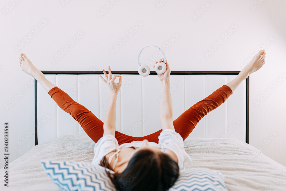 Naklejka premium Woman in bed while quarantine doing exercises in headphones, doing sports at home while self-isolation, home yoga, white bed and blue pillows.