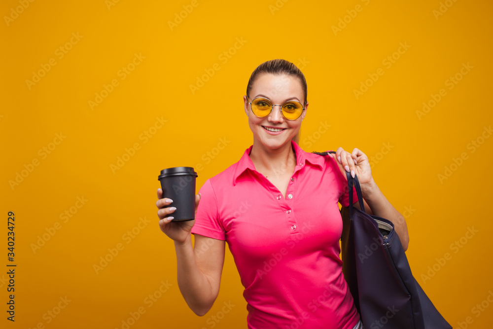 Cute blonde in sunglasses and t-shirt with bag on shoulder, drinking her hot drink, portrait on yellow background