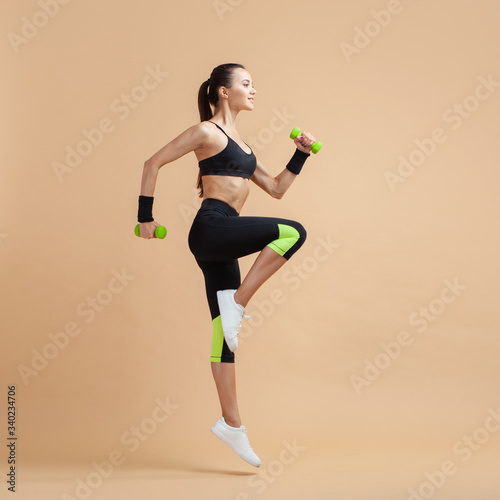 A young brunette woman is engaged in fitness, jumping with dumbbells, raising her knees high, on a peach background.