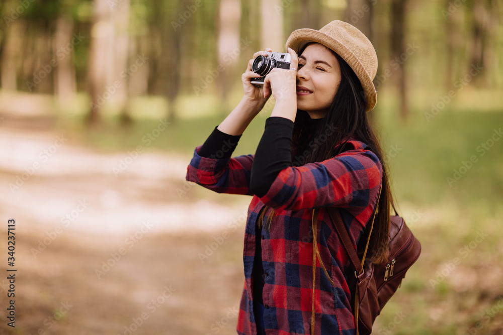 stylish hipster young woman  taking photos in the forest, carrying a backpack in the forest on sunset light in the spring season, looking at amazing woods, travel concept
