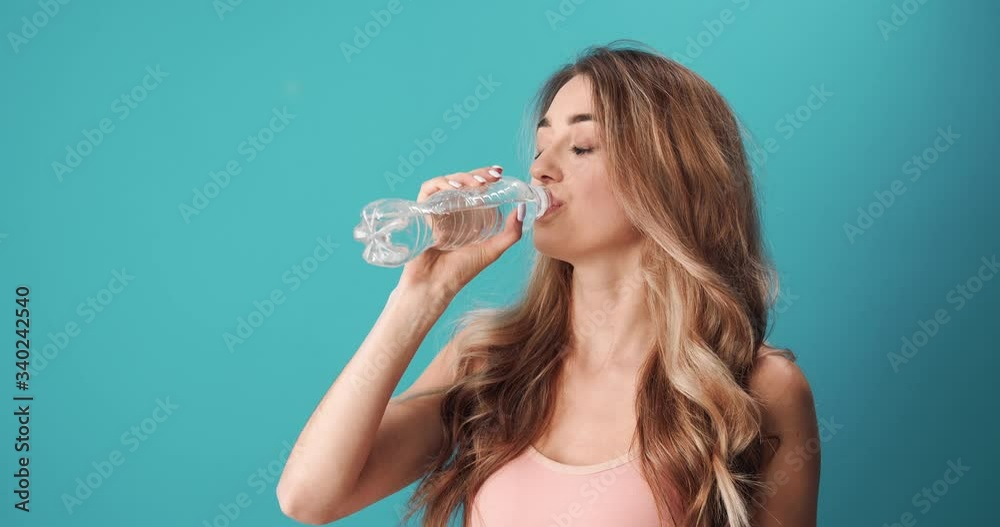Happy beautiful girl with long curly hair drinking water while standing in studio with blue background. Sporty woman refreshing after workout. Happy beautiful girl with long curly hair drinking water while standing in studio with blue background. Sporty woman refreshing after workout.
