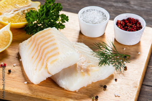 Fresh raw cod with seasonings and vegetables served on cutting board on wooden table
