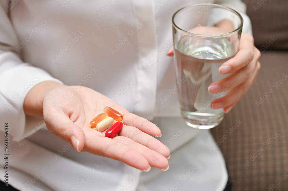 Close up Asian woman holding pill and a glass of water sitting on sofa. Concept of taking daily medicine, Multivitamins and supplements and medicine to cure head ache, stomach pain sedation meds. Close up Asian woman holding pill and a glass of water sitting on sofa. Concept of taking daily medicine, Multivitamins and supplements and medicine to cure head ache, stomach pain sedation meds.