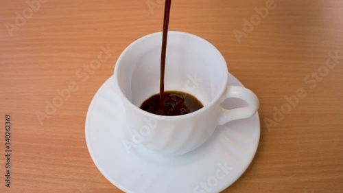 Slow motion coffee pour into a white porcelain cup on wooden table, with small plate. In natural light. Empty space for the copy.