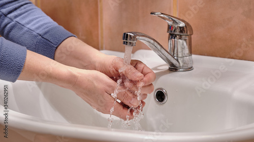 woman washes her hands under a stream of clear water from a chrome tap  white sink and terracotta background  stop covid-19 concept