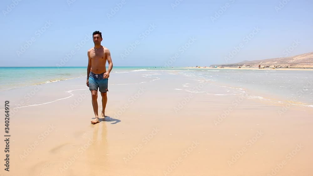 Young man walking away and turns around at empty Sotavento beach in Fuerteventura, Spain. Lonely male tourist enjoying summer vacation at seashore in Canary Islands. Holidays destination concept Young man walking away and turns around at empty Sotavento beach in Fuerteventura, Spain. Lonely male tourist enjoying summer vacation at seashore in Canary Islands. Holidays destination concept