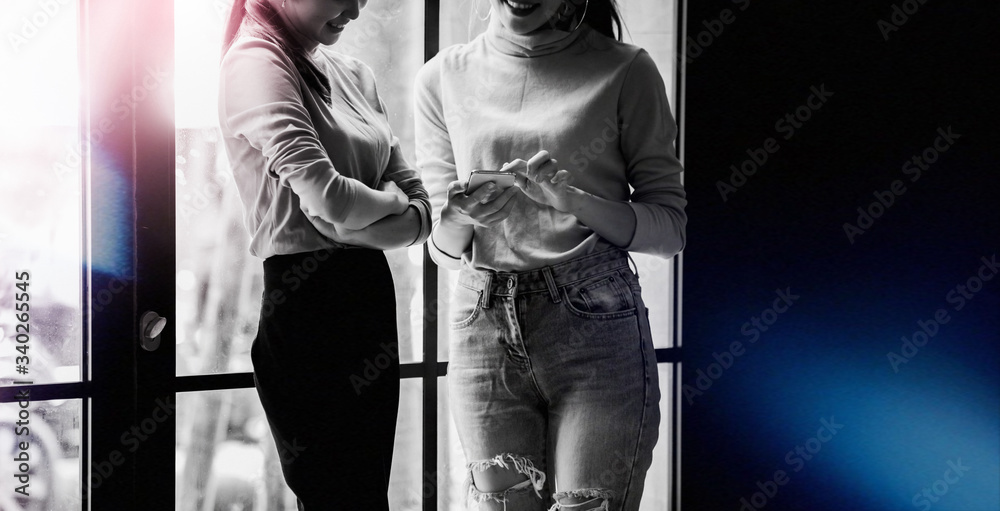 Two Charming Asian business women with the smartphone standing on a window. Girl shows her friend image on screen of smartphone. Young people problems with cell phone addiction