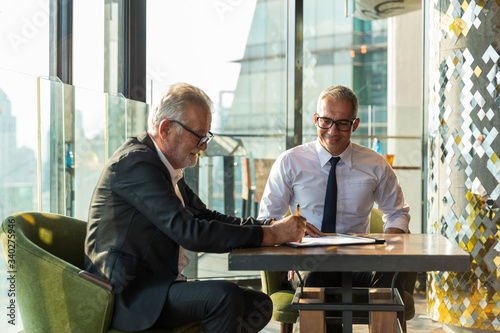 Picture of two business man discussing business on a table.  The young man is wearing a white shire with black tie and glass. The older man wear a black suit and sunglass. They are in a hotel lobby.