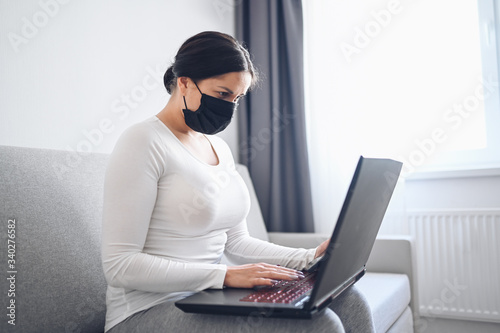 Young freelance woman in face medicine mask working on a laptop and phone on grey couch during coronavirus isolation home quarantine. Covid-19 pandemic Corona virus. Online work from home concept.