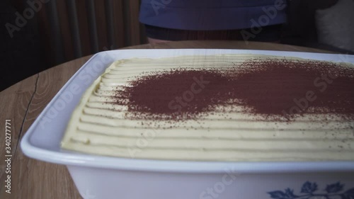 Homemade tiramisu, woman sprinkles cocoa powder on tiramisu. White plate with golden edges. Italian dessert on black kitchen background, close up photo