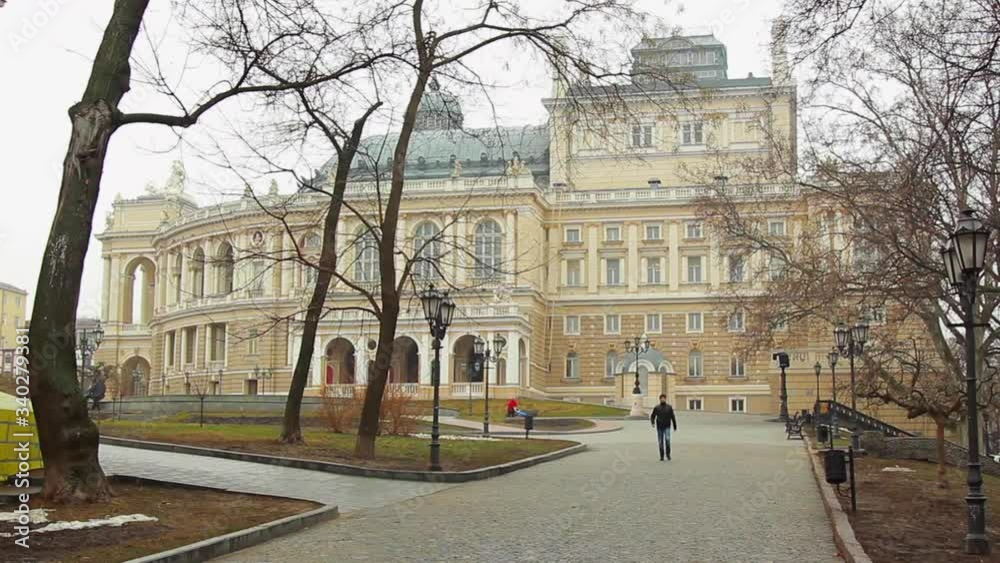 Odessa Opera and Ballet Theater in Odessa, Ukraine on a white sky background. Citizens walk along the paved walkways surrounded by leafless brown trees, wrought iron street lights and benches on a clo Odessa Opera and Ballet Theater in Odessa, Ukraine on a white sky background. Citizens walk along the paved walkways surrounded by leafless brown trees, wrought iron street lights and benches on a clo