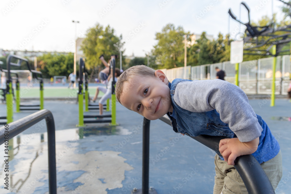 Happy small boy hanging on brachiating bar at a school yard. Other kids playing on levers for training in background. Selective focus. Happy small boy hanging on brachiating bar at a school yard. Other kids playing on levers for training in background. Selective focus.