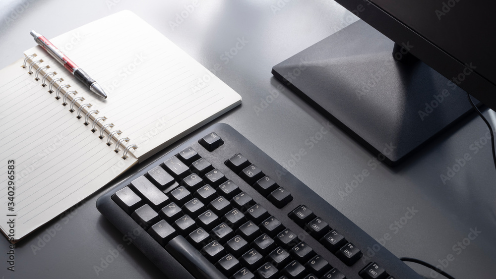 Selective focus at black keyboard with part of computer monitor, ballpoint pen and spiral note pad on gray tabletop with sunlight and shadow on surface Selective focus at black keyboard with part of computer monitor, ballpoint pen and spiral note pad on gray tabletop with sunlight and shadow on surface