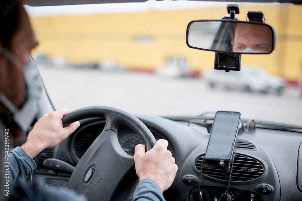 A male taxi driver in a medical mask gets into the car and turns on the Navigator on his smartphone