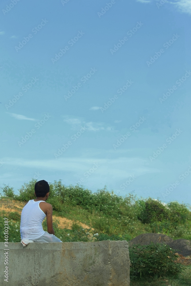 a teenager boy sitting roadside wearing traditional bengal dress dhoti and looking at the nature and blue sky