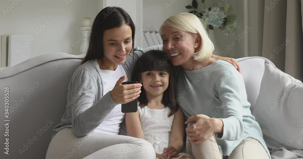 Excited little cute girl sitting between young mother and middle aged granny, using smartphone apps at home. Happy three generations female family enjoying free weekend time with electronic gadget. Excited little cute girl sitting between young mother and middle aged granny, using smartphone apps at home. Happy three generations female family enjoying free weekend time with electronic gadget.