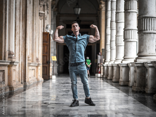 Full length strong young male in casual clothes showing biceps while standing in spacious hallway of old building