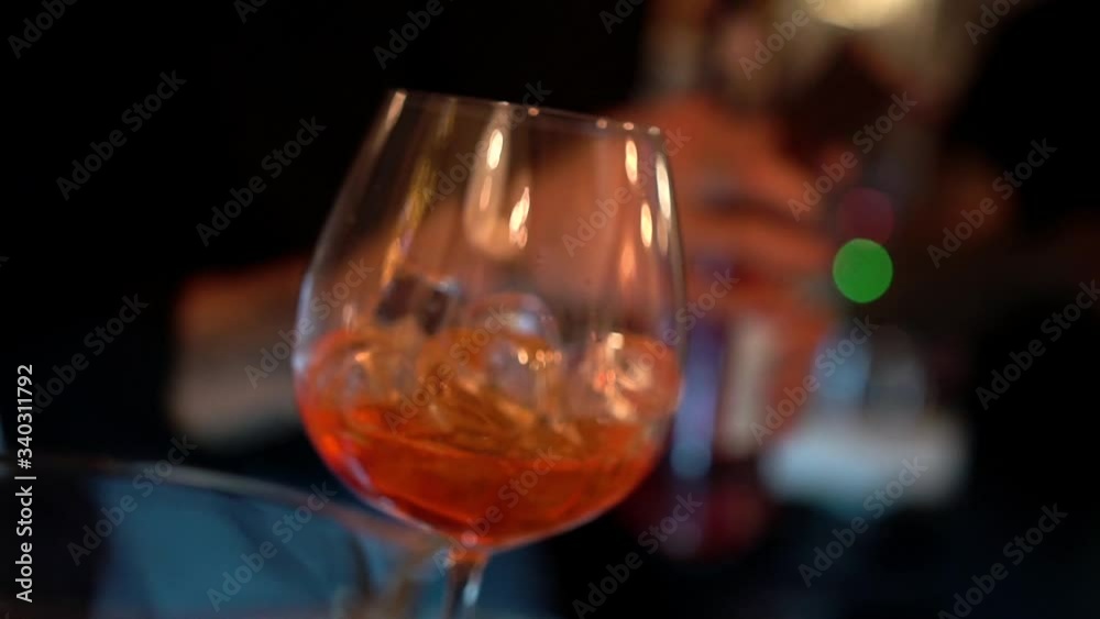 A bartender silhouette fills a transparent glass goblet with reflections with ice cubes and orange-colored alcohol drink on a bar background, close-up. Slow motion with blur bokeh lights.