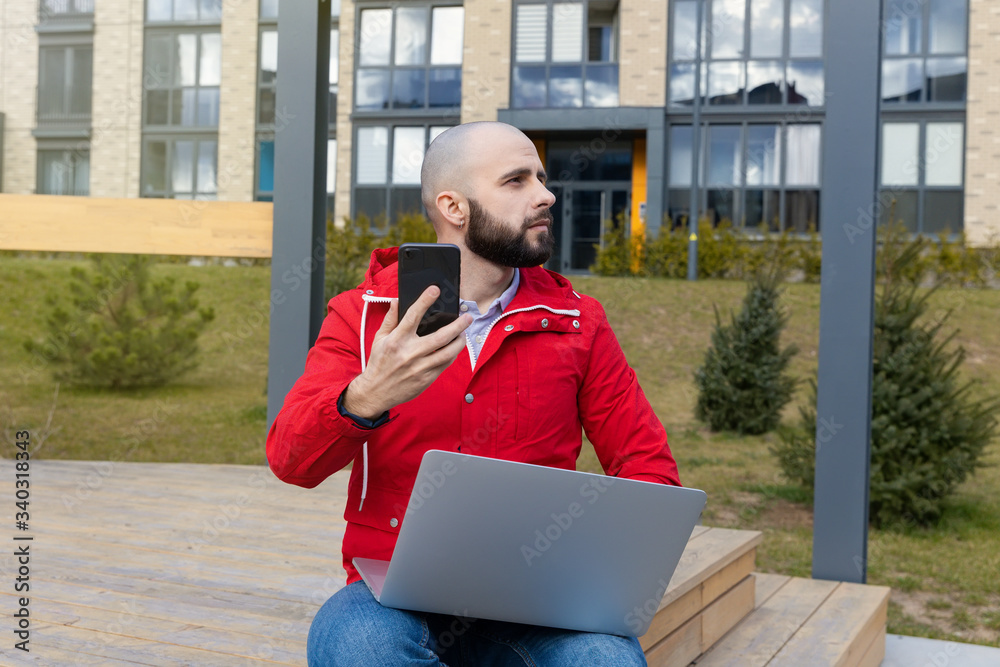 A brutal guy with a beard in everyday clothes works at a computer on the street. Concept of working outside the home A brutal guy with a beard in everyday clothes works at a computer on the street. Concept of working outside the home