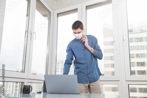 A guy in a protective face mask is talking on the phone and working at a computer from home during a virus epidemic. Quarantine during viral infection