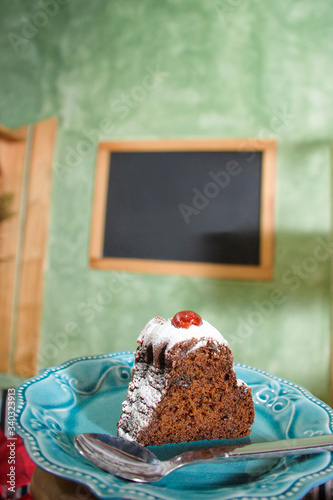 Piece of black cake covered in snow sugar and cherry served on a blue plate. In the background a frame