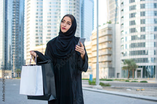 Photography Arab woman in Abaya holding cell phone and shopping bags on the city street