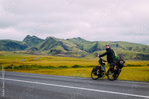 male cyclist traveler stands next to a bicycle and backpacks against the backdrop of mountains in Iceland