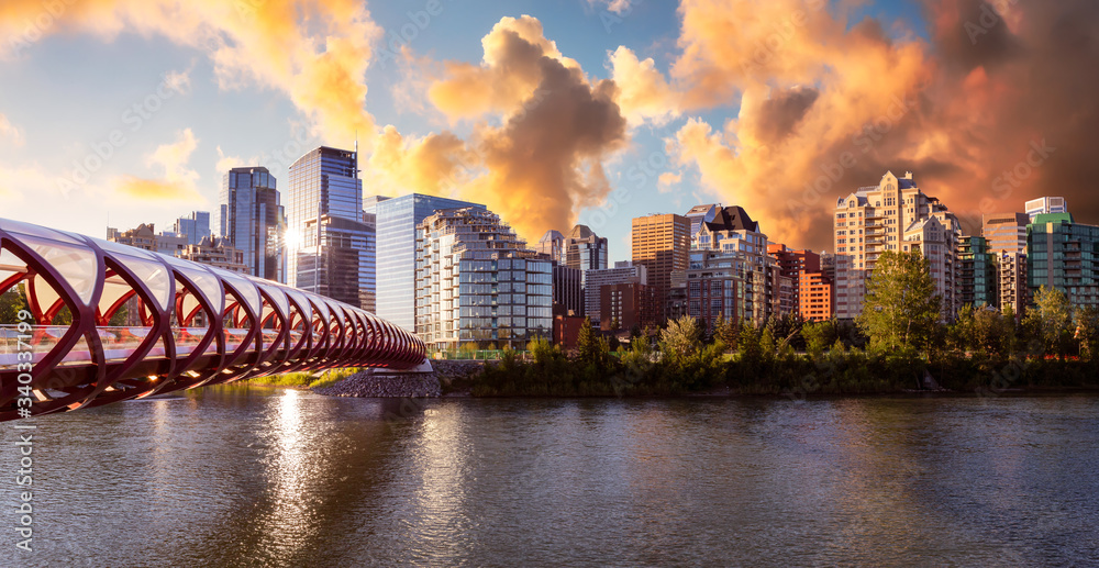 Fototapeta premium Peace Bridge across Bow River with Modern City Buildings in Background during a vibrant summer sunrise. Cloudy Sky Composite. Taken in Calgary, Alberta, Canada.