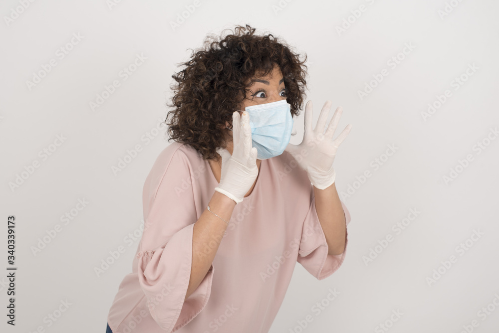 Portrait of young woman wearing medical mask with shocked facial expression holding hands near face, screaming and looking sideways at something amazing. Stands against gray studio Wall. Portrait of young woman wearing medical mask with shocked facial expression holding hands near face, screaming and looking sideways at something amazing. Stands against gray studio Wall.