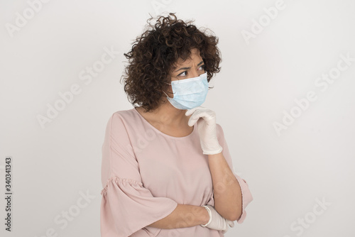Young European woman wearing medical mask with hand under chin and looking sideways with doubtful and skeptical expression, suspect and doubt. Standing indoors over background. © Jihan
