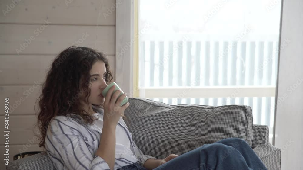 a curly-haired girl in jeans and a shirt drinks coffee from a birch mug in a beautiful white house against a large window. a curly-haired girl in jeans and a shirt drinks coffee from a birch mug in a beautiful white house against a large window.