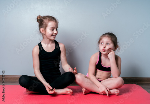Two girls in sports uniforms are sitting on a karemat and engaged in sports exercises at home during quarantine. Girls have fun.