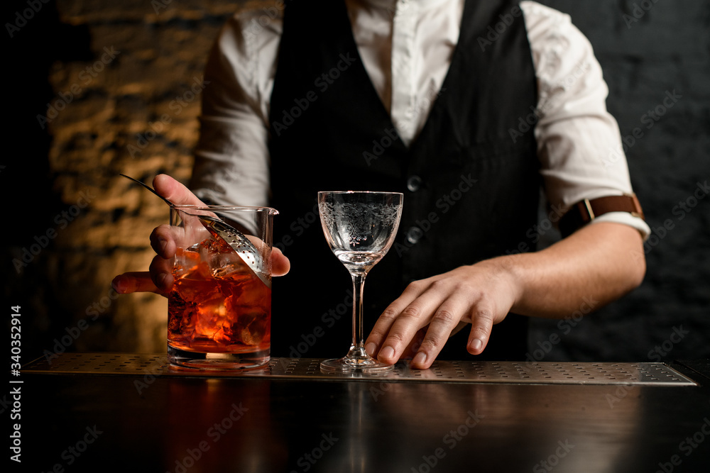 Close-up barman holds glassy mixing cup with strainer full of cold alcoholic drink with pieces of ice.