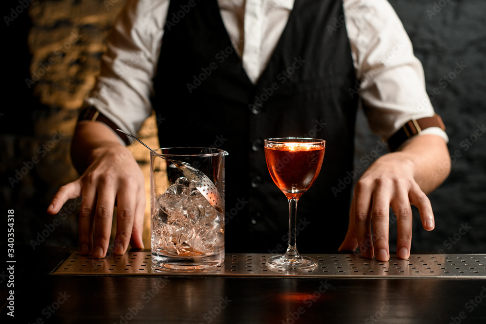 © fesenko - Close-up. Clear glassy mixing cup with strainer and glass full of cold alcoholic drink stand on bar