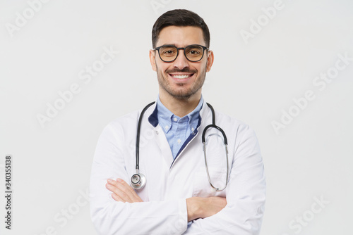 Close-up portrait of smiling handsome young male doctor with stethoscope around neck, wearing white coat and eyeglasses, isolated on gray background