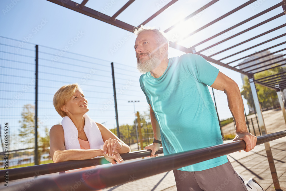 Senior family working out at outdoor gym. Mature bearded man in sportswear doing push ups on parallel bars. Active senior couple exercising in the morning