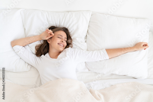 Young female dressed in white, lying on big bed, spreading arms wide, stretching to wake better, keeping eyes closed but smiling