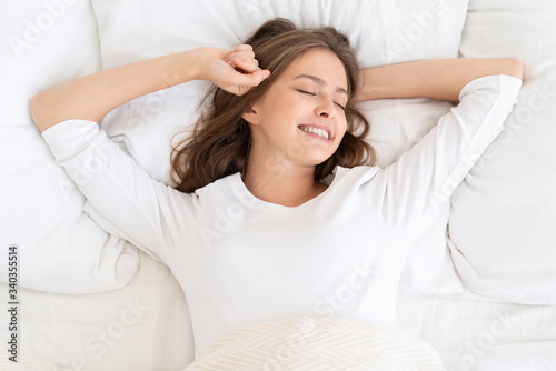 Young woman lying on bed late in morning, spending free time in bed with closed eyes, stretching elbows to wake up, showing lazy sleepy smile